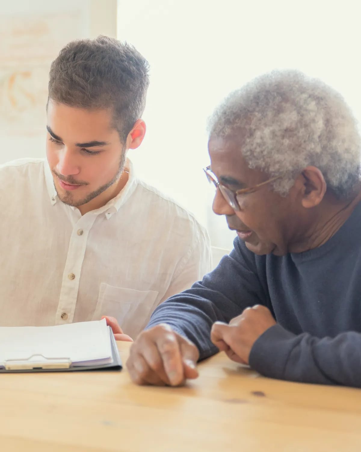 A young man and an older man go over papers on a clipboard at a wooden table