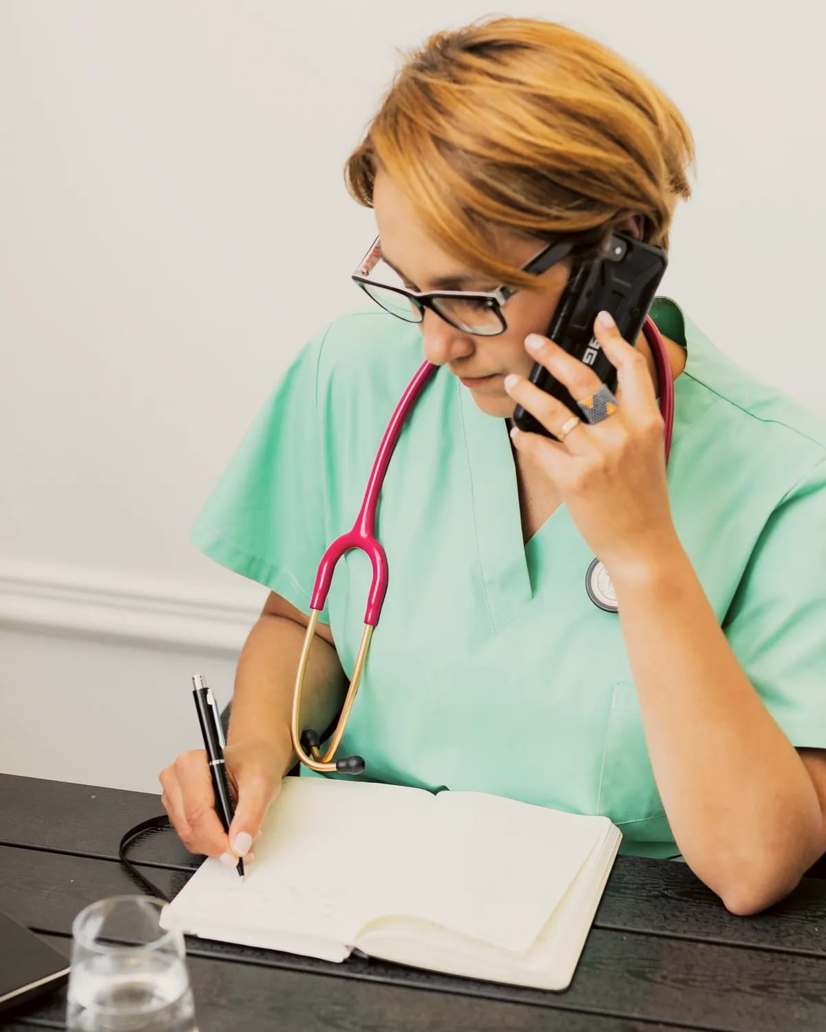 A nurse in green scrubs writes in a notebook while she talks on the phone