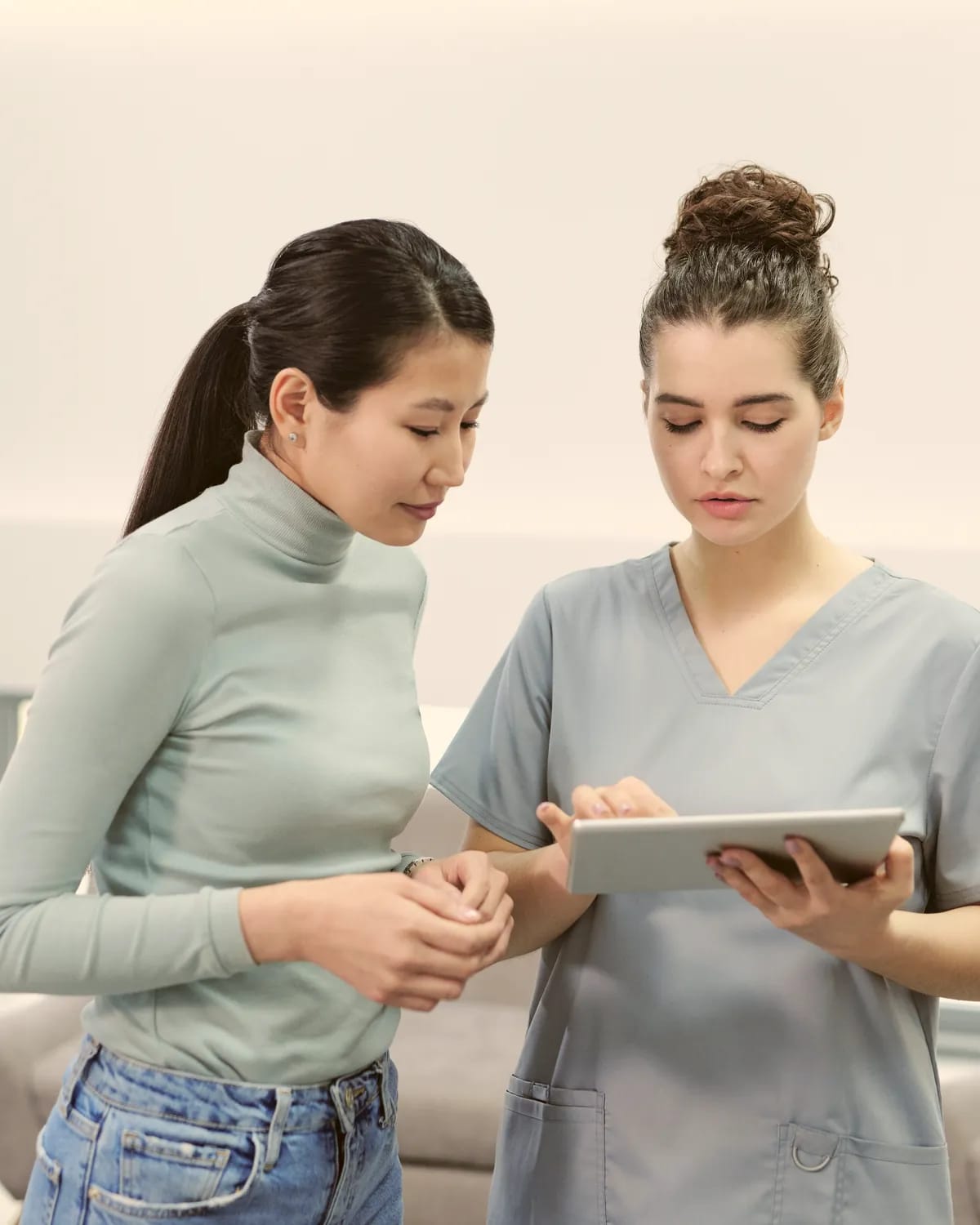 A nurse in scrubs and another woman look at a tablet together in a bright room